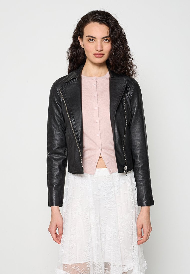 Frau mit lockigem Haar, die eine schwarze Lederjacke über einem rosa, geknöpften Oberteil und einem weißen Spitzenrock trägt, steht vor einem schlichten hellen Hintergrund.