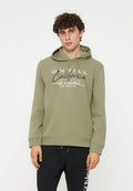 Junger Mann mit lockigem Haar, der einen olivgrünen Hoodie mit Text und schwarze Hosen vor einem schlichten weißen Hintergrund trägt.