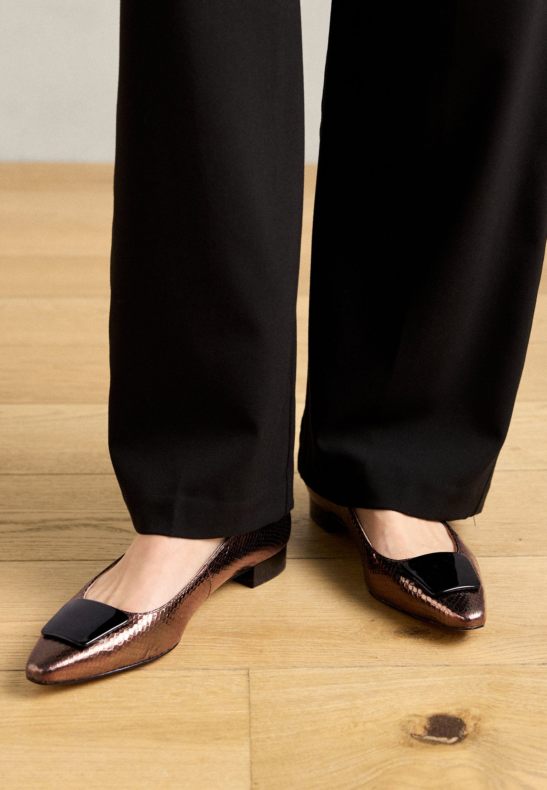 Bronzefarbene Ballerinas mit einer strukturierten, metallischen Oberfläche, die an der Spitze einen schwarzen, glänzenden Akzent aufweisen. Niedriger Blockabsatz, spitze Form.