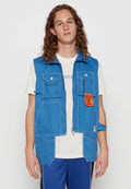 Blaues Jeansweste mit Reißverschluss, mehreren Taschen und einem orangefarbenen Aufnäher. Getragen über einem weißen Hemd, das kontrastierende Nähte aufweist.
