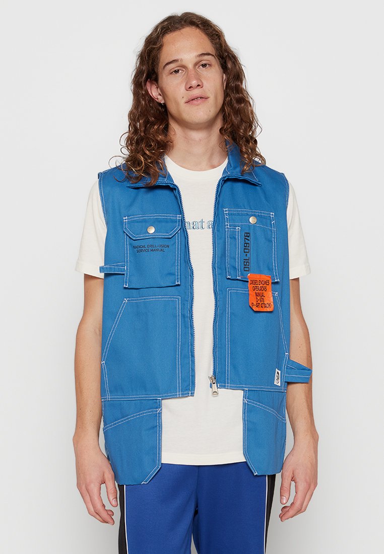 Blaues Jeansweste mit Reißverschluss, mehreren Taschen und einem orangefarbenen Aufnäher. Getragen über einem weißen Hemd, das kontrastierende Nähte aufweist.