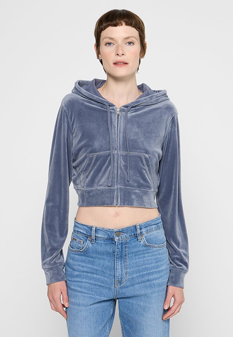 Kurze Kapuzenjacke aus hellgrauem Samt mit Reißverschluss, Kapuze und zwei vorderen Taschen. Kombiniert mit hochtaillierten blauen Jeans.