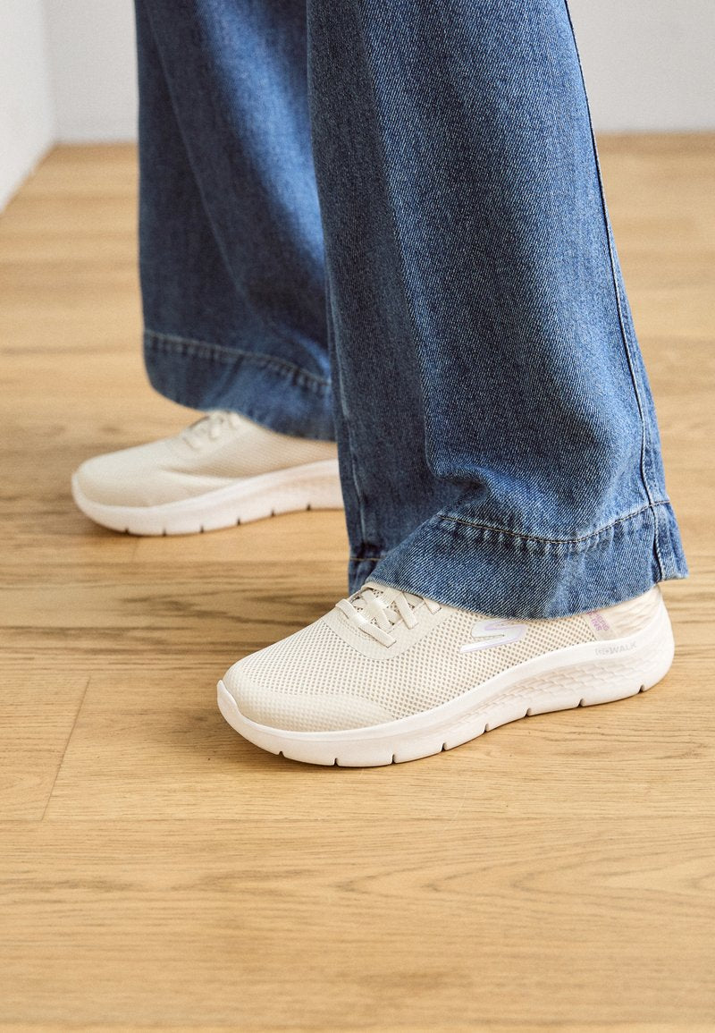 Leichte, beige Sportschuhe mit einem Mesh-Obermaterial, flexibler Sohle und Schnürdesign, kombiniert mit weitgeschnittenen blauen Jeans.