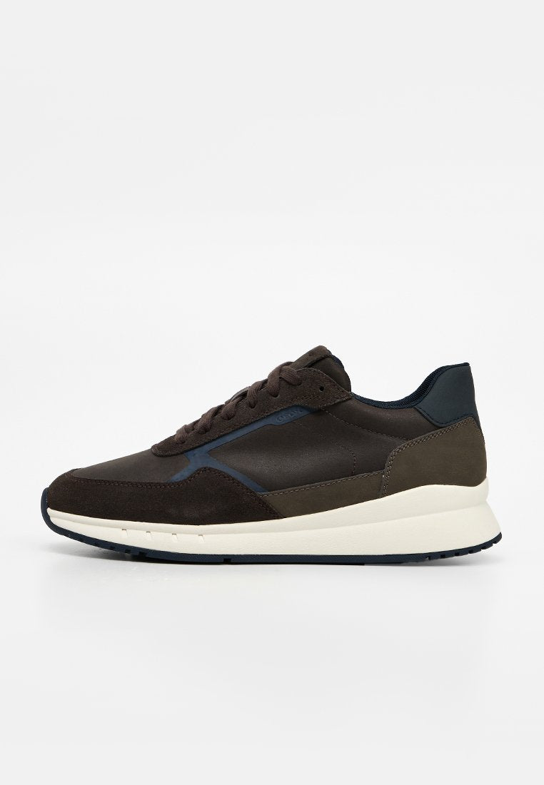 Dunkelbrauner niedriger Sneaker mit Obermaterial aus Wildleder und Leder, weißer dicker Sohle und marineblauem Kragen, von der Seite auf weißem Hintergrund gezeigt.