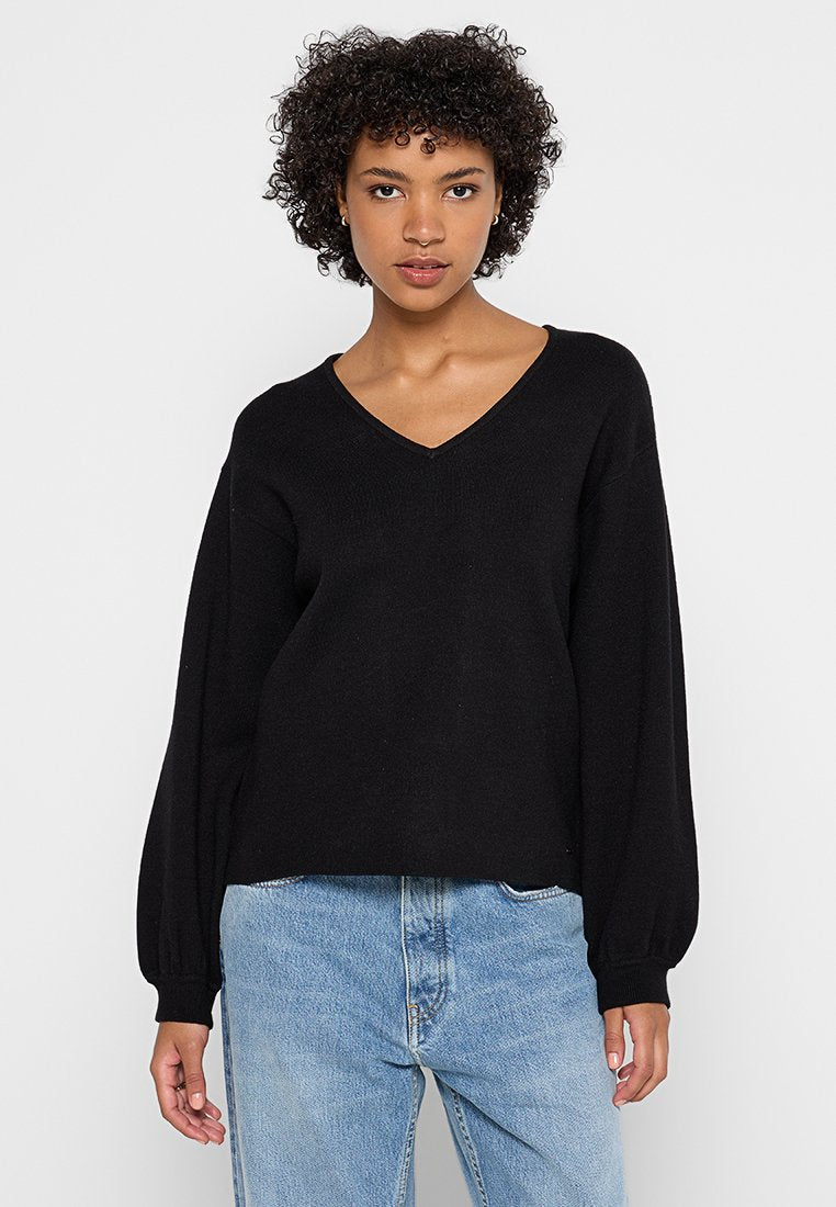 Junge Frau mit lockigem Haar, die einen schwarzen V-Ausschnitt-Pullover mit weit ausgestellten Ärmeln und hellblauen, hochgeschnittenen Jeans trägt, vor einem schlichten Hintergrund.
