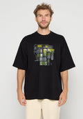 Mann trägt ein oversized schwarzes T-Shirt mit Grafik verschiedener vintage Handys in grünen und grauen Tönen auf der Vorderseite.