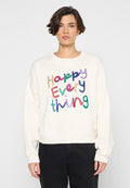 Frau trägt weißen Pullover mit farbig glitzerndem Text "Happy Everything" und schwarze Hose, steht vor einfachem hellem Hintergrund.