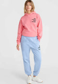 Rosa Kapuzenpullover mit schwarzem Text und hellblauen Jogginghosen, beide aus weichem Material. Weiße Sneakers runden das Outfit ab.