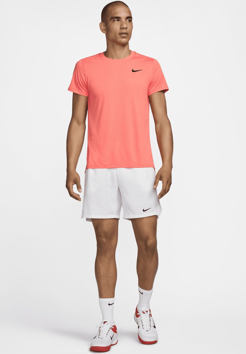Nike Sportoutfit mit einem hellkorallfarbenen T-Shirt mit kurzen Ärmeln, weißen Shorts, weißen Socken und sportschuhen mit roten Akzenten.