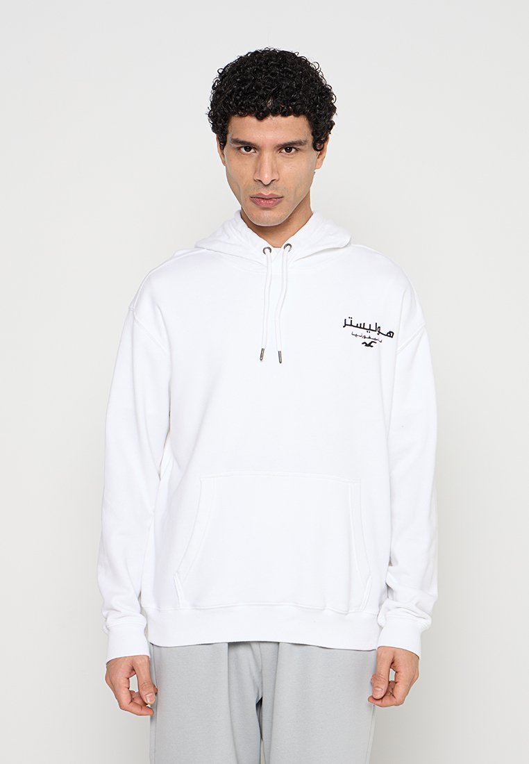 Mann trägt weißen Hoodie mit schwarzem arabischem Text und kleinem Logo auf der Brust, steht vor schlichtem hellem Hintergrund.