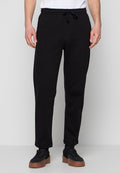 Mann trägt schwarze Sweatpants mit Kordelzug und schwarze Sneakers mit braunen Sohlen, steht vor einem schlichten hellgrauen Hintergrund.