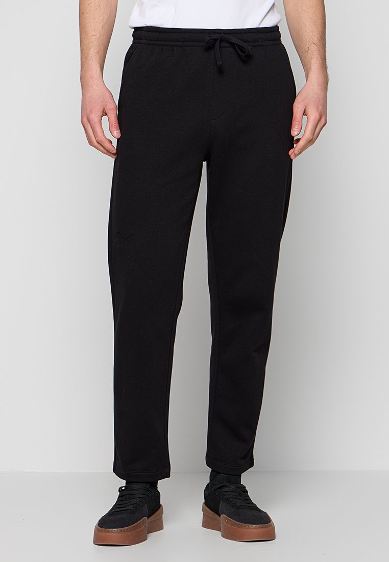 Mann trägt schwarze Sweatpants mit Kordelzug und schwarze Sneakers mit braunen Sohlen, steht vor einem schlichten hellgrauen Hintergrund.