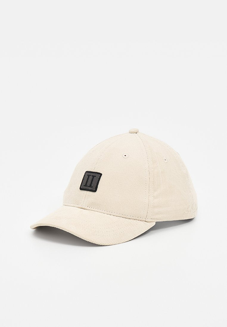 Beige Kordhut mit gebogenem Schirm und schwarzem rechteckigem Patch, auf dem zwei vertikale weiße Linien mittig auf der Vorderseite angeordnet sind.
