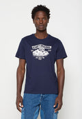 Mann trägt ein dunkelblaues T-Shirt mit einem weißen Motorrad und dem Text ""Schott Original Speed, New York City 1913"", kombiniert mit blauen Jeans.