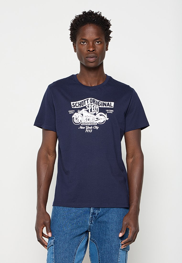 Mann trägt ein dunkelblaues T-Shirt mit einem weißen Motorrad und dem Text ""Schott Original Speed, New York City 1913"", kombiniert mit blauen Jeans.