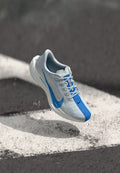 Weiße und blaue Nike ZoomX-Laufschuhe fliegen über schwarzen Asphalt mit einer weißen bemalten Linie und verstreuten Staubpartikeln.