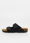 Schwarze Slides-Sandale mit zwei verstellbaren Riemen, Kork-Fußbett und schwarzer Gummisohle vor neutralem hellgrauem Hintergrund.