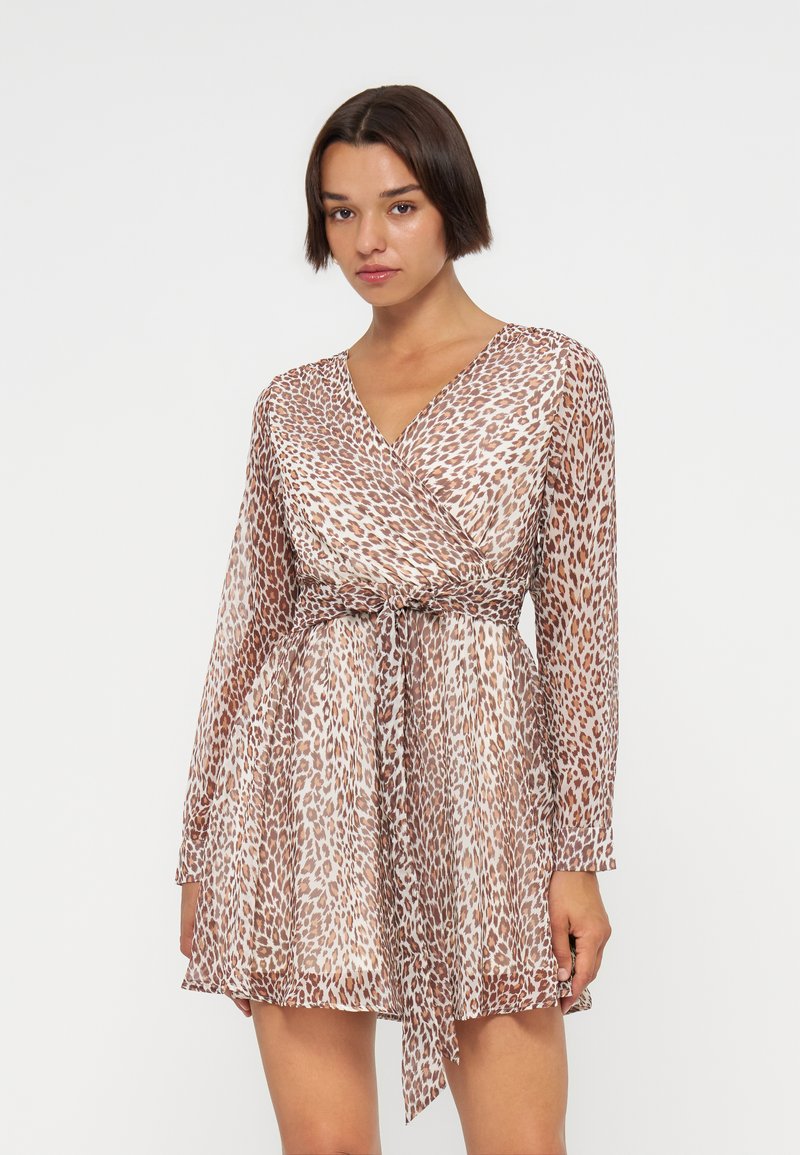 Junge Frau mit kurzen dunklen Haaren, die ein langärmeliges Wickelkleid mit Leopardenmuster und gebundener Taille trägt und vor einem schlichten Hintergrund steht.