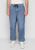 Mann trägt lockere blaue Jeans mit schwarzem Kordelzug, weiße Turnschuhe und weiße Socken, steht vor einem schlichten hellen Hintergrund.