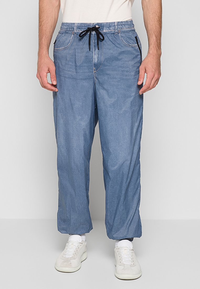 Mann trägt lockere blaue Jeans mit schwarzem Kordelzug, weiße Turnschuhe und weiße Socken, steht vor einem schlichten hellen Hintergrund.