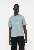 Mann trägt ein hellblau gesprenkeltes T-Shirt mit einem Smiley-Design und schwarze Shorts, steht vor einem einfarbigen weißen Hintergrund.