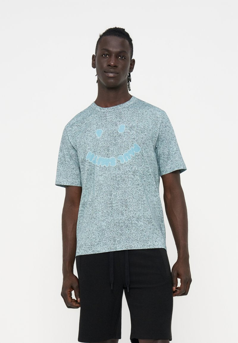 Mann trägt ein hellblau gesprenkeltes T-Shirt mit einem Smiley-Design und schwarze Shorts, steht vor einem einfarbigen weißen Hintergrund.