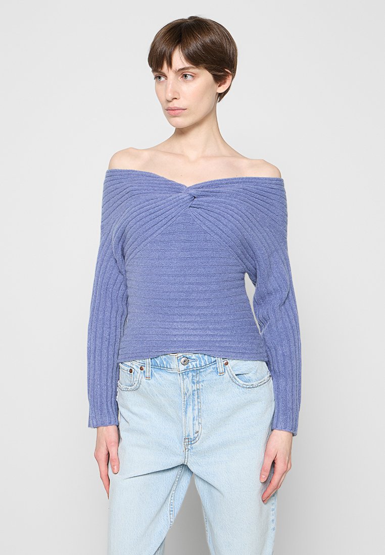 Frau mit kurzen braunen Haaren, die einen gerippten, schulterfreien Lavendelpullover und hellblaue, hoch taillierte Jeans trägt, steht vor einem einfarbigen Hintergrund.