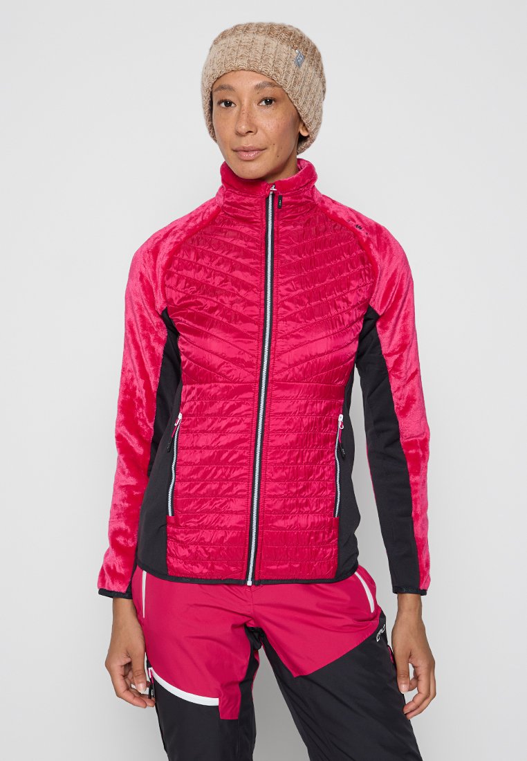 Frau trägt eine beigefarbene Strickmütze, eine rote gesteppte Jacke mit schwarzen Seitenpaneelen und passende rot-schwarze Outdoor-Hosen, steht vor einem weißen Hintergrund.