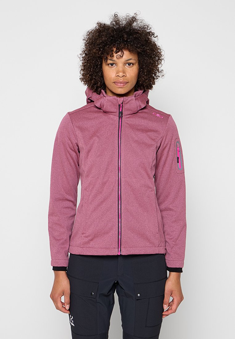 Frau mit rosa Softshelljacke mit Reißverschluss und Kapuze sowie schwarzen Cargohosen steht vor einem schlichten hellen Hintergrund.