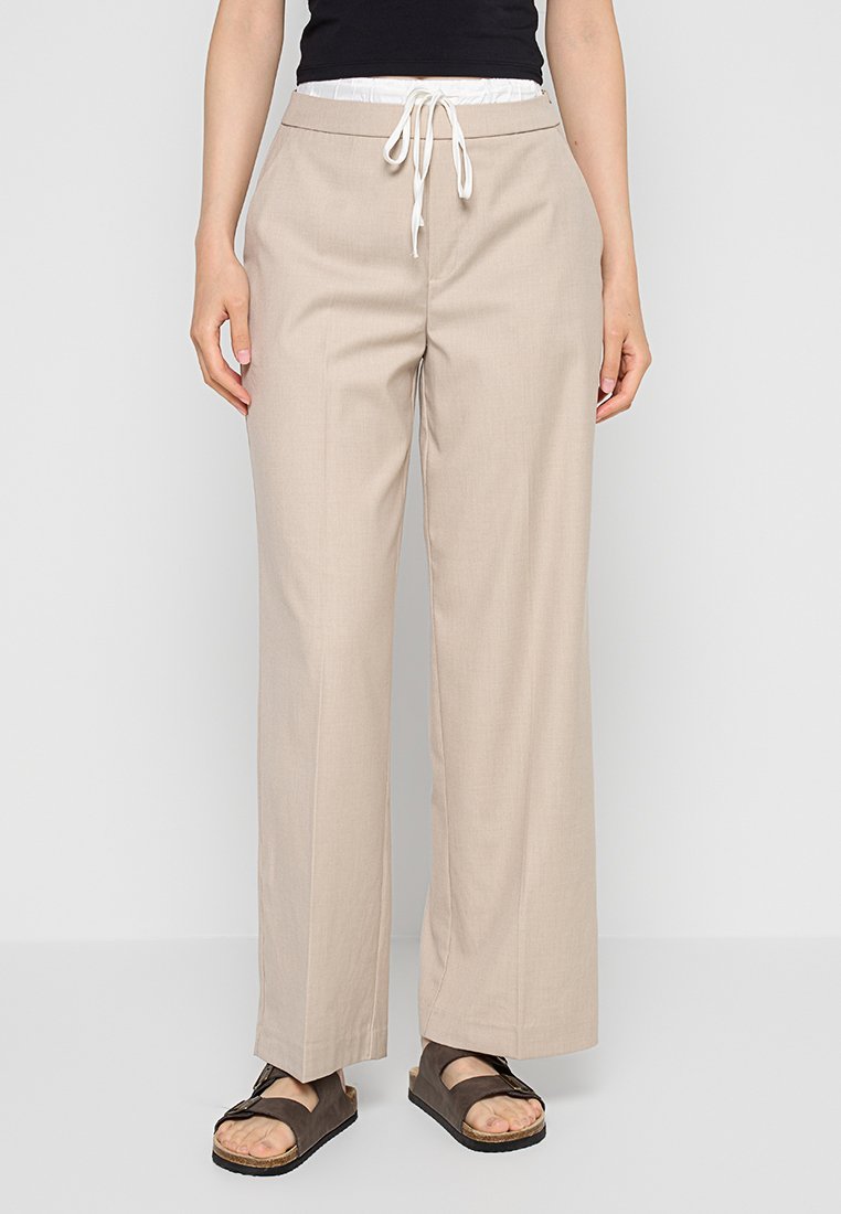 Beige weite Hosen mit einem Kordelzugbund und einer leichten Textur. Kombiniert mit dunklen Sandalen. Sichtbares Modell von der Taille abwärts.