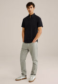 Schwarzes Kurzarm-Hemd mit Knöpfen, graue Hose und weiße Sneaker mit braunen Akzenten. Das Modell steht mit Händen in den Taschen vor einem neutralen Hintergrund.