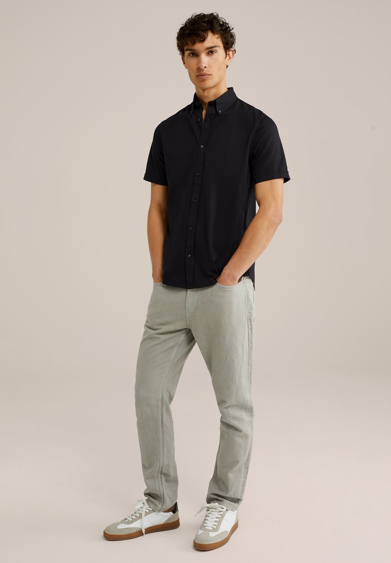 Schwarzes Kurzarm-Hemd mit Knöpfen, graue Hose und weiße Sneaker mit braunen Akzenten. Das Modell steht mit Händen in den Taschen vor einem neutralen Hintergrund.
