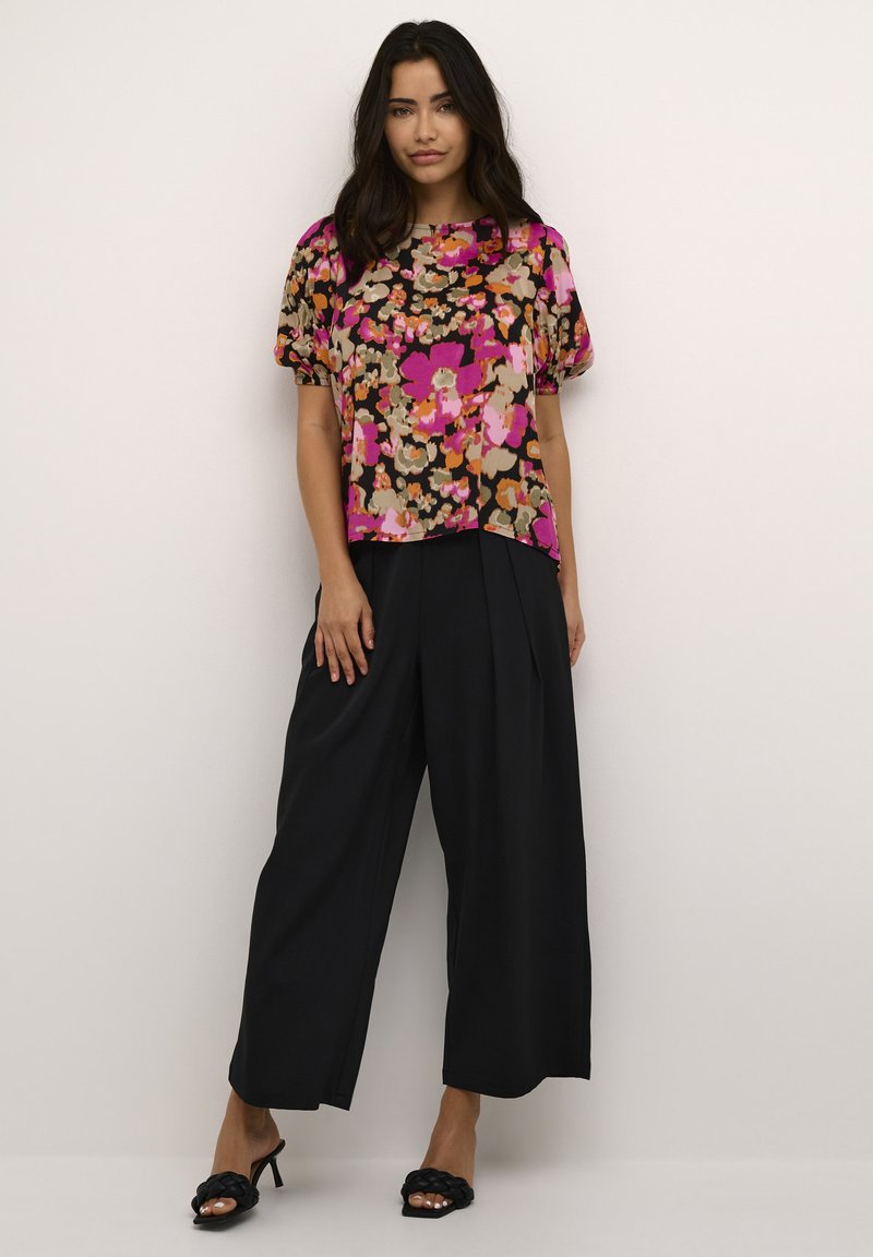 Bluse mit kurzen Ärmeln im Blumenmuster auf schwarzem Hintergrund, mit rosa, braunen und cremefarbenen Blumen. Kombiniert mit weit geschnittenen schwarzen Hosen und schwarzen High Heels.