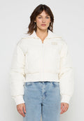 Frau trägt eine weiße, kurze Steppjacke mit gerippten Bündchen und Kragen, kombiniert mit hellblauen, hoch taillierten Jeans, stehend vor weißem Hintergrund.