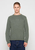 Mann trägt grünen Tommy Jeans Strickpullover und blaue Jeans, steht vor einem schlichten hellgrauen Hintergrund.