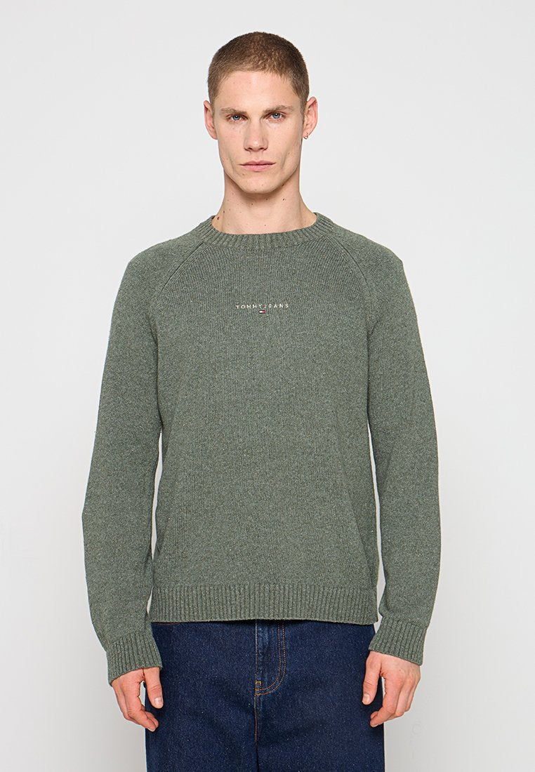 Mann trägt grünen Tommy Jeans Strickpullover und blaue Jeans, steht vor einem schlichten hellgrauen Hintergrund.