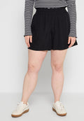 Person trägt schwarze Shorts mit elastischem Bund, schwarz-weiß gestreiftes Langarmshirt und weiße Sneakers, steht vor einfachem Hintergrund.