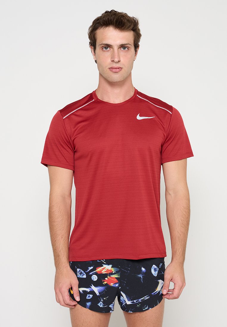 Junger Mann, der ein rotes Nike-T-Shirt und schwarze Shorts mit abstraktem Muster trägt, steht vor einem weißen Hintergrund.