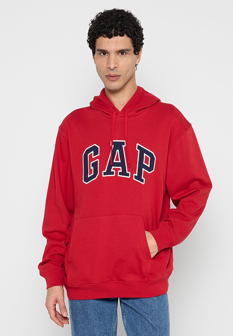 Junger Mann mit lockigem dunklem Haar, der einen roten GAP-Hoodie und blaue Jeans trägt, steht mit einer Hand in der Bauchtasche.