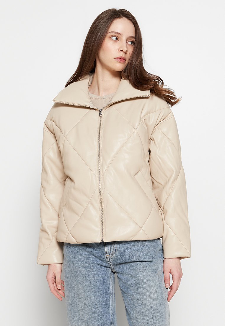 Beige Kunstlederjacke mit diamantförmigem Steppmuster, hohem Kragen, Frontreißverschluss und lockerer Passform, kombiniert mit hellblauen Jeans.