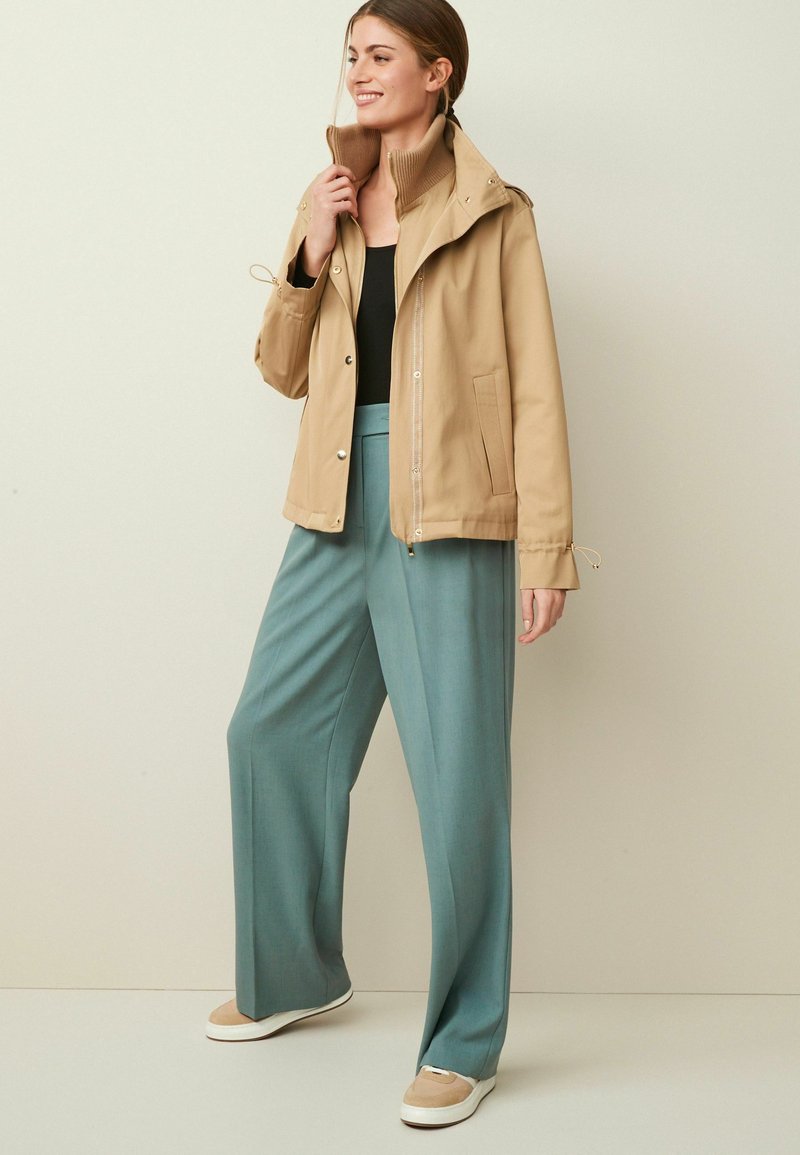 Frau lächelt, trägt eine beige Jacke über einem schwarzen Oberteil, türkisfarbene weite Hosen und beige Sneakers, steht vor einem schlichten hellen Hintergrund.