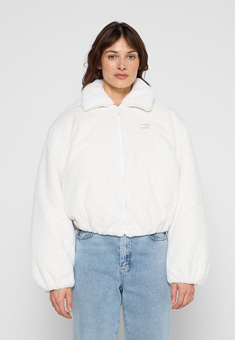 Frau trägt eine weiße, kuschelige Jacke mit Reißverschluss und elastischen Bündchen sowie hellblaue, hoch taillierte Jeans, steht vor einem schlichten Hintergrund.