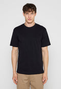 Junger Mann steht frontal, trägt ein schlichtes schwarzes T-Shirt mit kurzen Ärmeln und beige Hosen vor einem grauen Hintergrund.