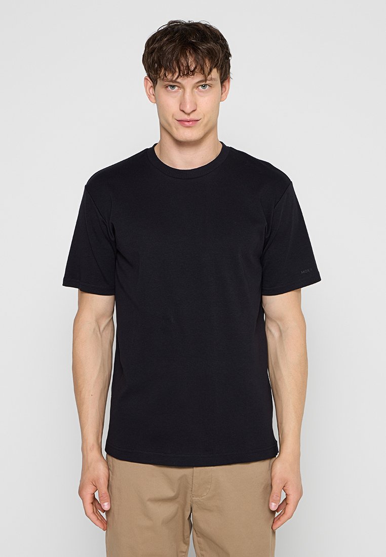 Junger Mann steht frontal, trägt ein schlichtes schwarzes T-Shirt mit kurzen Ärmeln und beige Hosen vor einem grauen Hintergrund.