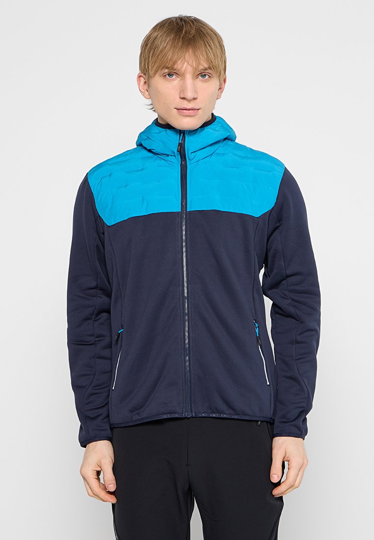 Junge Person in einer marineblauen und leuchtend blauen Zip-Jacke mit Kapuze, die vor einem schlichten hellen Hintergrund steht.