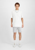 Weißes, übergroßes Baumwoll-T-Shirt, graue Shorts mit Logo und weiße, mehrfarbige Sneakers. Das Model steht vor einem schlichten Hintergrund.