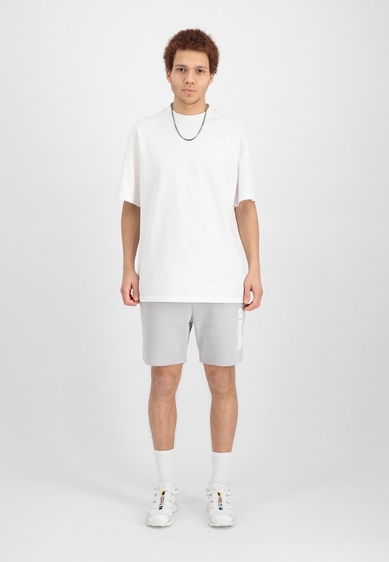 Weißes, übergroßes Baumwoll-T-Shirt, graue Shorts mit Logo und weiße, mehrfarbige Sneakers. Das Model steht vor einem schlichten Hintergrund.