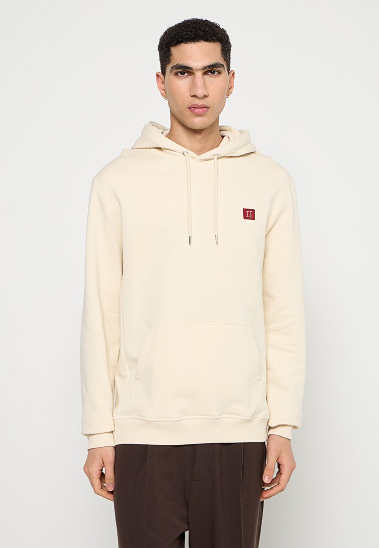 Beige Kapuzenpullover mit einer Fronttasche, Kordelzugkapuze und kleinem roten Logo auf der linken Brust. Hergestellt aus weichem Stoff mit glatter Textur.