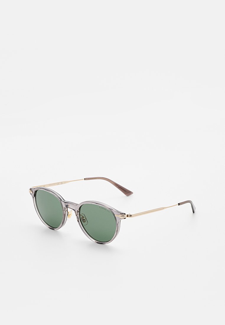 Runde Sonnenbrille mit grünen Gläsern, durchscheinendem grauem Kunststoffrahmen und dünnen goldfarbenen Metallbügeln mit braunen Enden, auf weißem Hintergrund dargestellt.