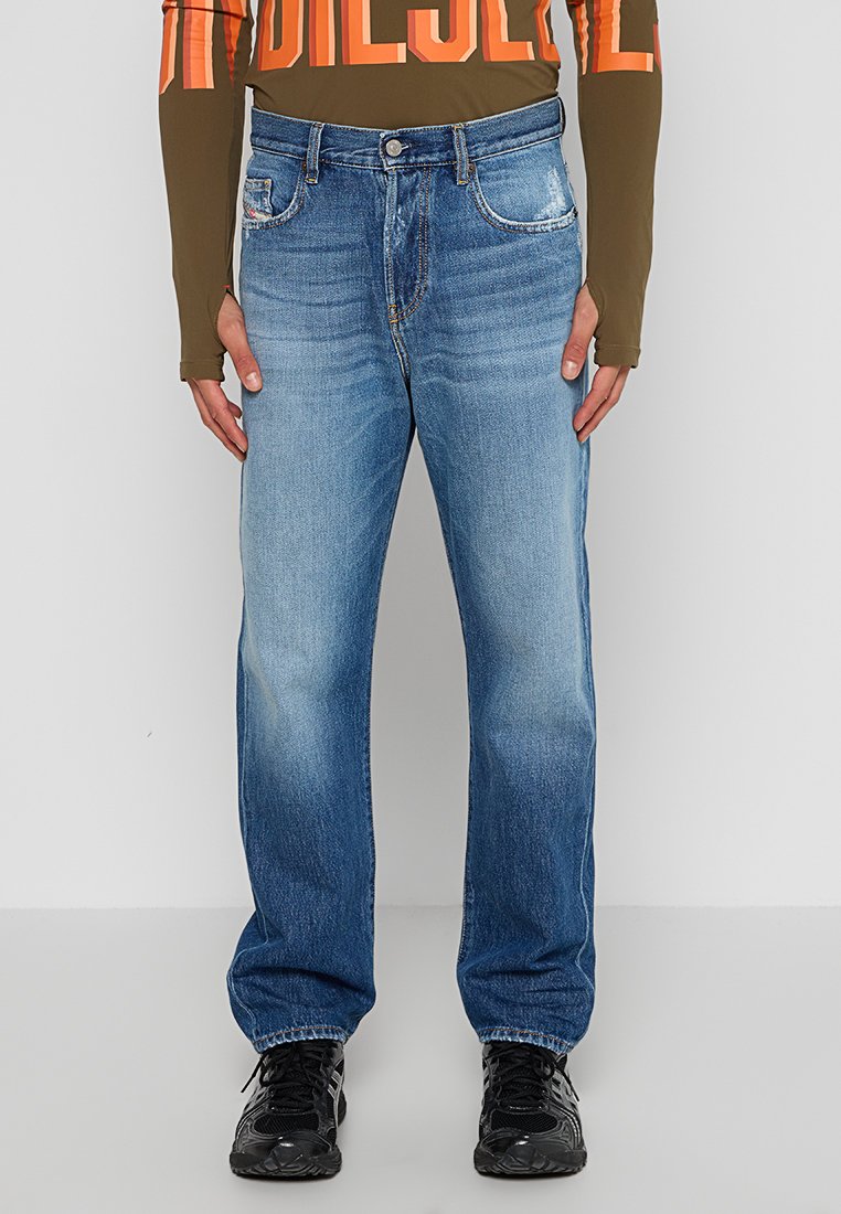 Blaue Straight-Leg-Jeans mit ausgewaschenen Stellen, fünf Taschen und Knopfverschluss, kombiniert mit einem braunen Langarmoberteil und schwarzen Sneakers.